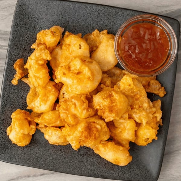 Golden-fried battered shrimp or chicken pieces on a dark plate with a side of sweet chili sauce.